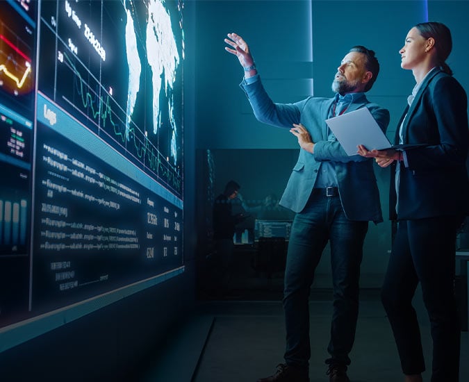 Two people gesturing to a map and data on a large video wall. Two people gesturing to a map and data on a large video wall.