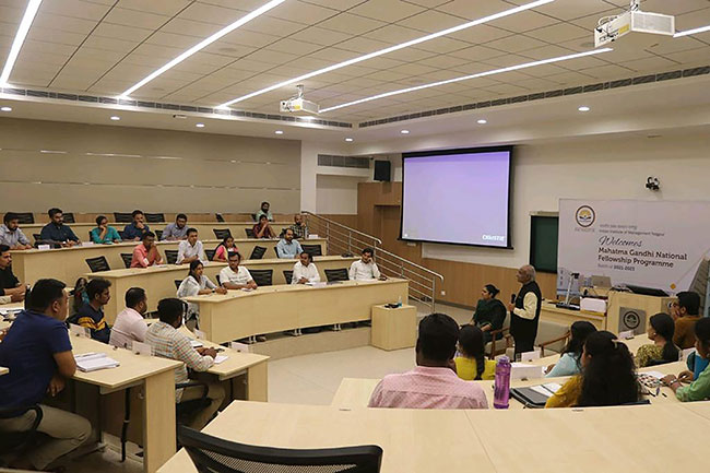 Students in a classroom in India sitting in tiered seating listening to a lecture, with a screen on the wall in the background, and two Christie projectors hanging from the ceiling.