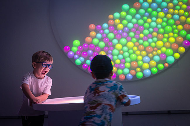 Two children standing around a table with an image of colorful balls in the background. 