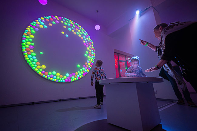 A woman and child stand around a table. The woman is pointing to a projected image of balls on the wall.