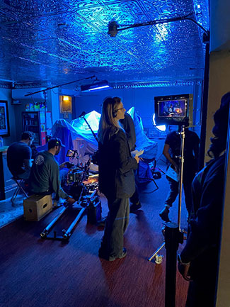 A woman stands looking at a film monitor with members of a film crew in the background.  