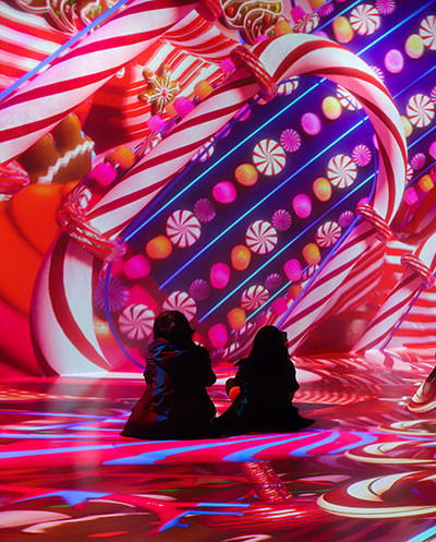 Two kids look at images of candy canes, gum drops, and gingerbread cookies projection mapped onto a wall and floor.