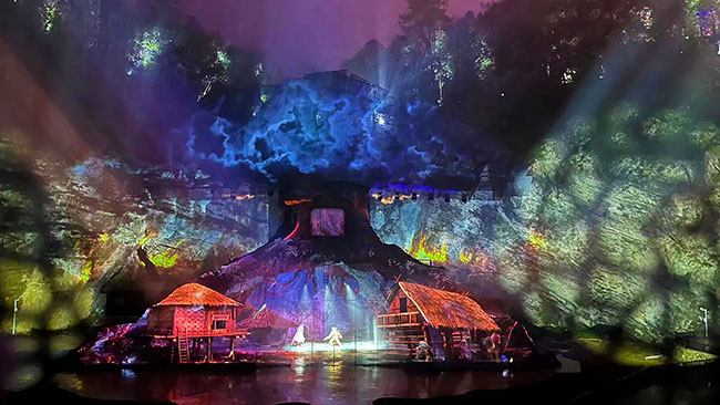 Performers illuminated by red and yellow lights on a stage with a tree as a backdrop.