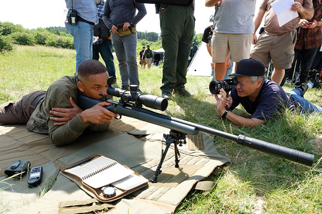 Man lying on the ground looking through the scope of a gun while another lies diagonal to him taking pictures.