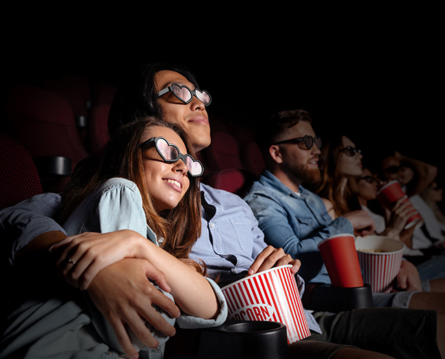 A couple in a theatre watching a movie while wearing heart-shaped glasses. 