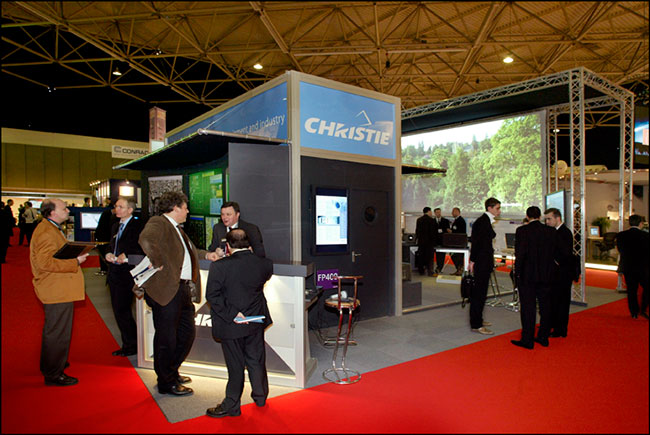 A tradeshow booth with a large screen in the background and people talking in groups.