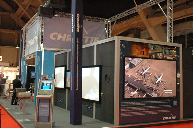 A tradeshow booth with the word “Christie” shown throughout and three large screens on the booth walls.