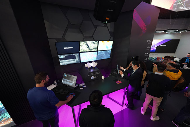 People stand around a desk with two computer monitors, with a three by two array of LCD panels in the background. 