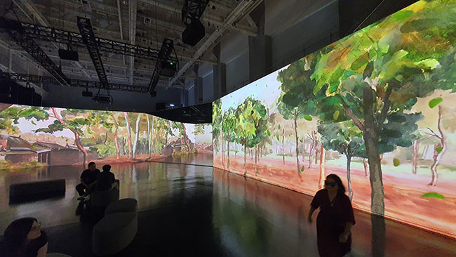 People in a large, darkened room with images of painted trees projected onto the walls.