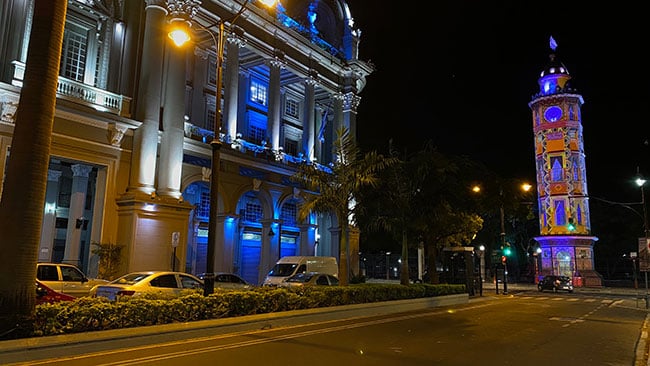 A street view of a building and clock tower. The clock tower is projection mapped with intricate designs.