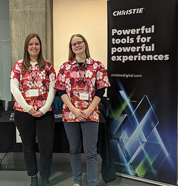 Two women wearing matching red and pink patterned shirts pose in front of a table. 