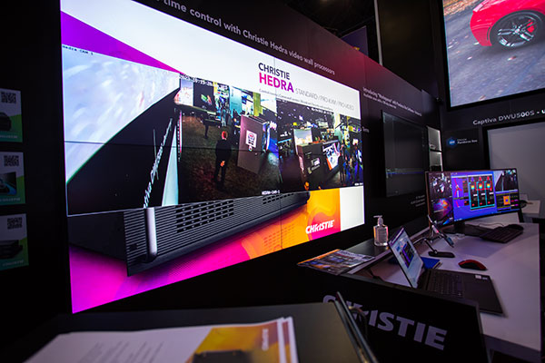 A video wall mounted on a black wall displays live camera feeds of people standing in a trade show booth.