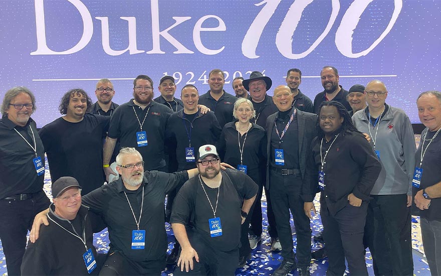 A group of people stand in front of a large sign with the text, “Duke 100.”