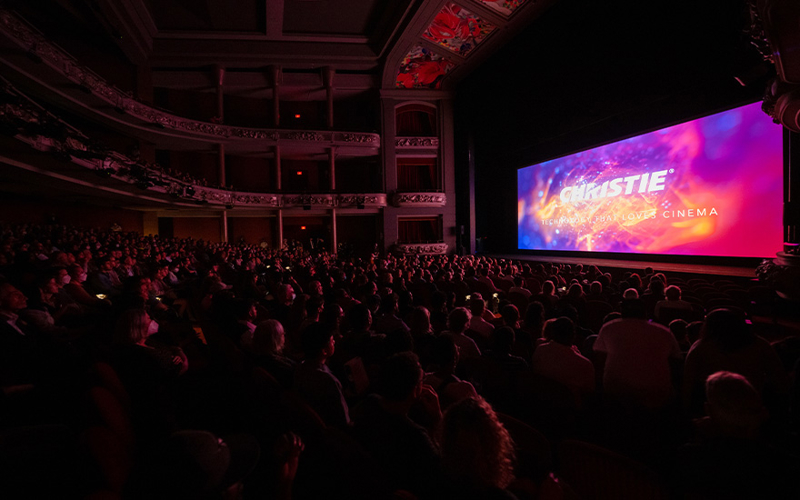 An audience in a large movie theatre facing a screen with the Christie logo on it
