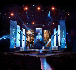 A man talking from a large stage with vertical screens on either side of him projecting images